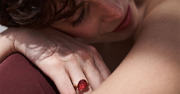 Persona con los ojos cerrados, apoyada en un hombro; lleva un gran anillo con una gema roja. Iluminación tenue.